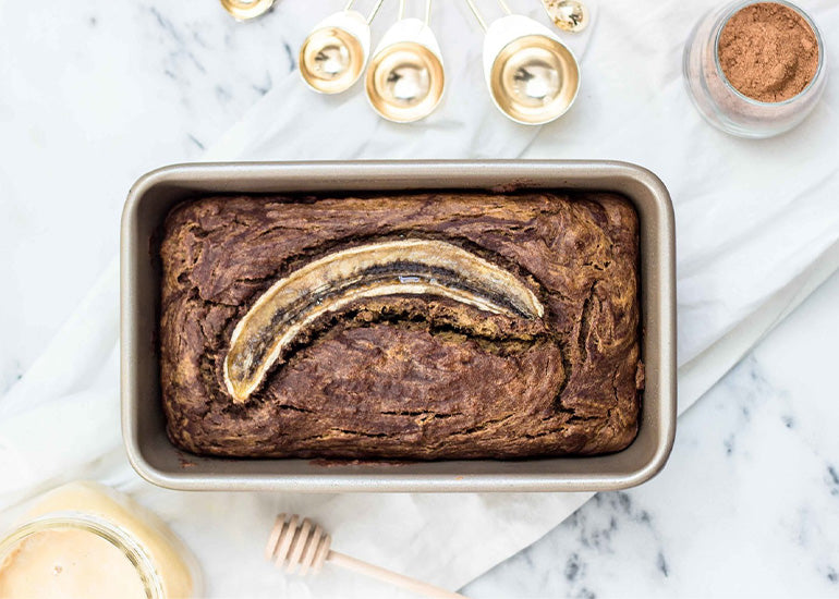 A baking pan with a loaf of chocolate banana bread made with Navitas Organics Cacao & Greens Essential Blend and Cacao Powder, topped with a sliced banana