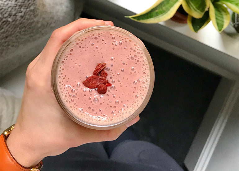 A woman holding a pink smoothie made with Navitas Organics Vanilla & Greens Essential Blend, Goji Powder and Chia Seeds, topped with Navitas Organics Goji Berries