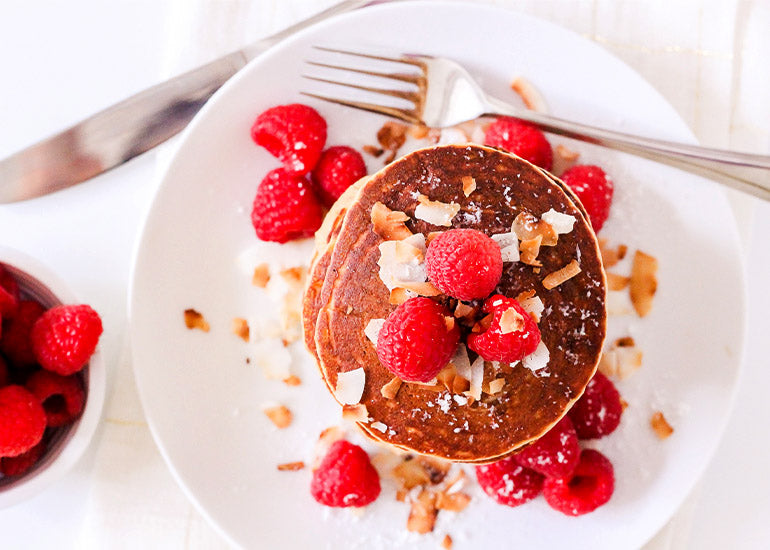 A stack of almond coconut paleo pancakes made with Navitas Organics Gelatinized Maca Powder, topped with maple syrup, coconut flakes and fresh raspberries