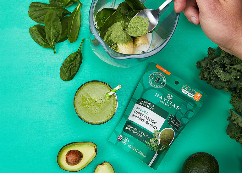 A green smoothie made with Navitas Organics Superfood+ Greens Blend, surrounded by avocados, kale and spinach