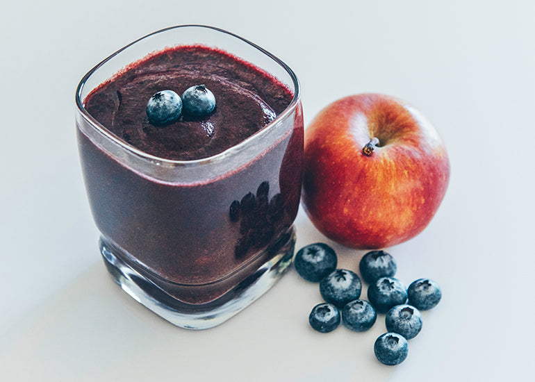 A smoothie made with Navitas Organics Acai Powder, Chia Seeds and Hemp Seeds in a glass next to fresh blueberries and an apple.