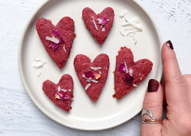 A woman holding a plate of heart-shaped cookies made with Navitas Organics Goji Berry Powder, topped with coconut flakes and rose petals.
