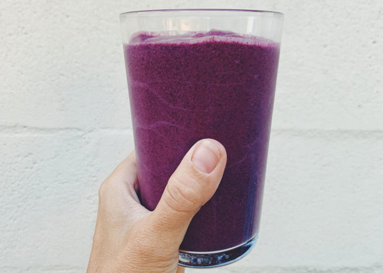 A woman holding a tall glass filled with a dark purple smoothie made with Navitas Organics Pomegranate Powder and Maqui Powder.