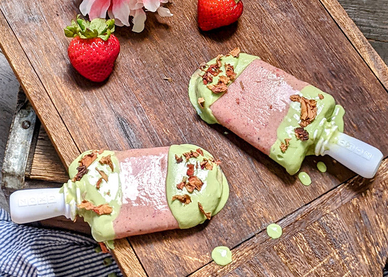 Two strawberry matcha popsicles made with Navitas Organics Superfood+ Berry Blend and Matcha Powder, on a wooden boars with strawberries and flowers.