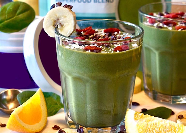 Two glasses filled with a green smoothie made with Navitas Organics Vanilla & Greens Essential Blend Protein Powder and Superfood+ Greens Blend, topped with Navitas Organics Goji Berries and Cacao Nibs.