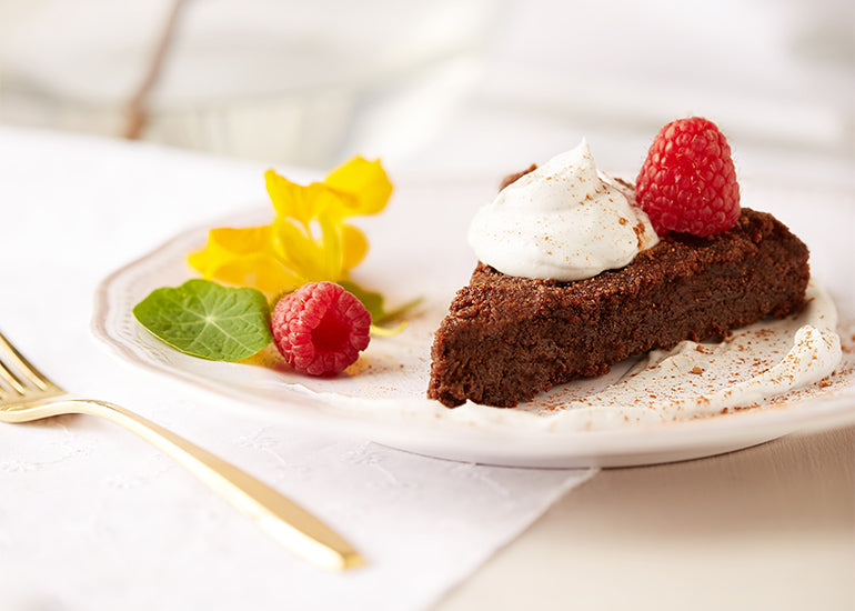 A slice of flourless chocolate cake made with Navitas Organics Cacao Powder and Maca Powder, topped with non-dairy whipped cream and a fresh raspberry.