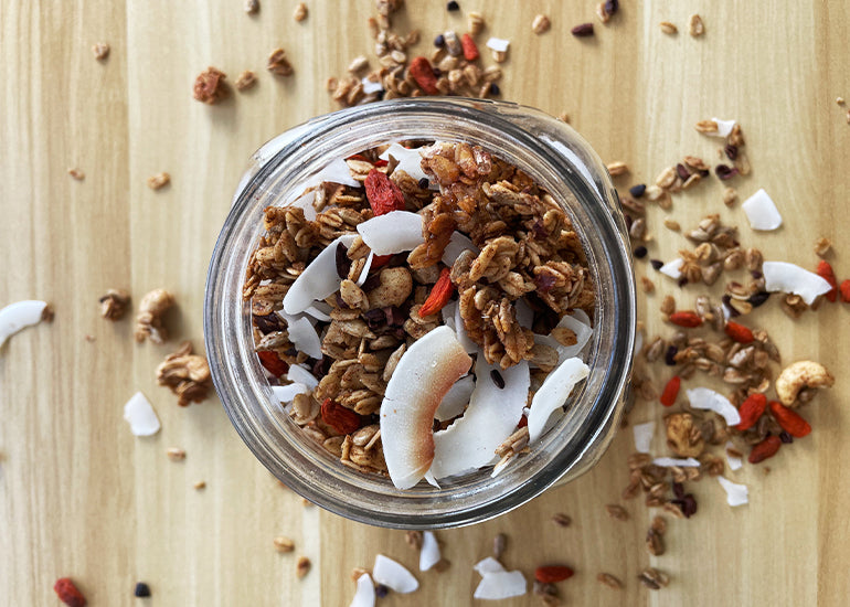 A mason jar filled with granola made with Navitas Organics Cashew Nuts and Maca Powder, topped with coconut flakes, and Navitas Organics Cacao Nibs and Goji Berries.