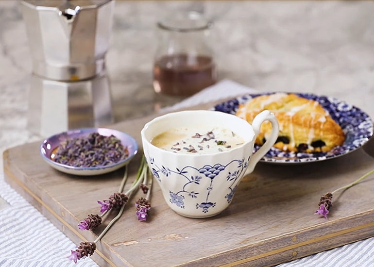 A porcelain cup filled with a lavender latte made with Navitas Organics Vanilla & Greens Essential Blend on a table with a scone and fresh lavender.