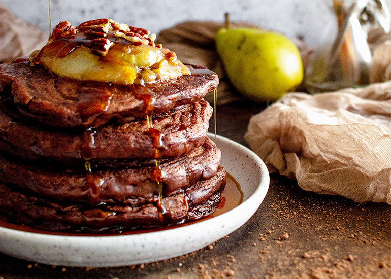 A stack of chocolate pancakes made with Navitas Organics Cacao Powder, drizzled with maple syrup and topped with pear compote and crushed pecans.