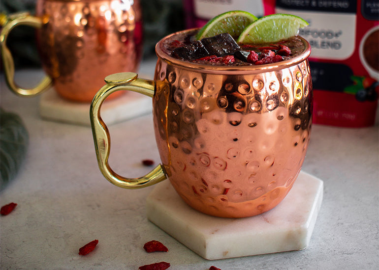 A superfood mocktail mule made with Navitas Organics Superfood+ Berry Blend served in a traditional Moscow mule mug topped with Navitas Organics Goji Berries and lime wedges.