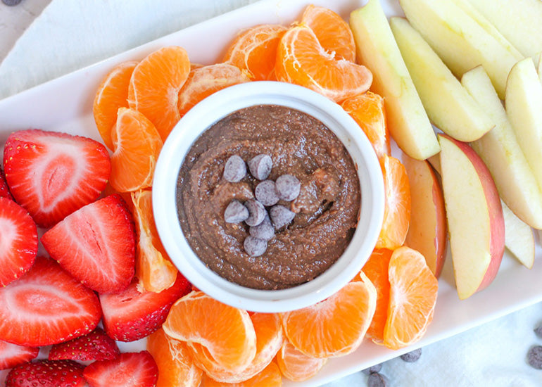 A dish filled with chocolate hummus made with Navitas Organics Cacao Powder and Cacao Butter, topped with chocolate chips, surrounded by fresh sliced fruit