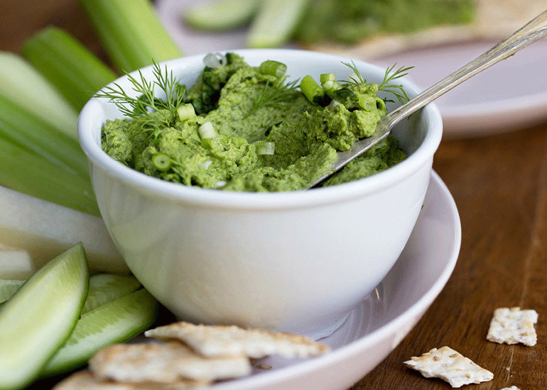 A bowl of green hummus made with Navitas Organics Wheatgrass Powder on a dish with celery sticks and crackers for dipping.
