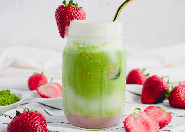 A mason jar filled with an iced strawberry matcha latte made with Navitas Organics Cacao Butter Wafers and Matcha Powder, topped with a vegan whipped cream and a fresh strawberry.