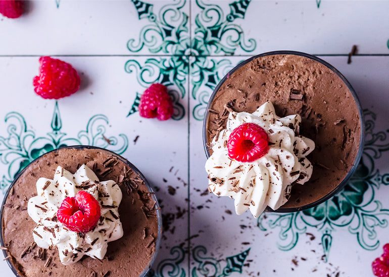 Two cups of chocolate raspberry maca mousse made with Navitas Organics Cacao Wafers, Maca Powder and Chia Seeds, topped with whipped cream, fresh raspberries and chocolate shavings.