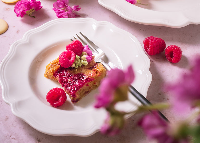 A white chocolate raspberry brownie made with Navitas Organics Cacao Butter Wafers on a plate topped with fresh raspberries.