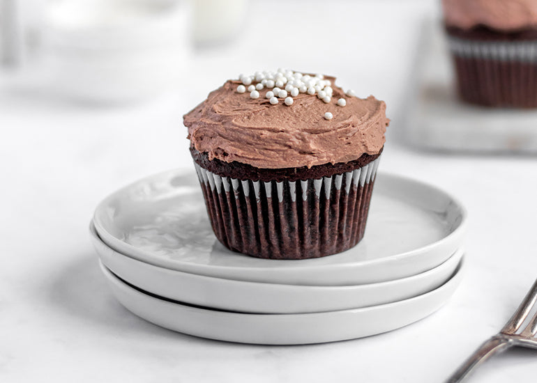 A chocolate cupcake made with Navitas Organics Cacao Powder and Cacao Wafers topped with whipped chocolate frosting and sprinkles sitting on a stack of white plates.