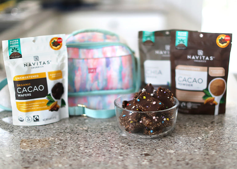 A dish filled with no-bake cacao bars made with Navitas Organics Unsweetened Cacao Wafers, Cacao Powder and Chia Seeds sitting on a countertop with a child's backpack.