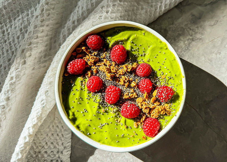 A creamy green smoothie bowl made with Navitas Organics Superfood+ Greens Blend, topped with fresh raspberries, gluten-free granola and Navitas Organics Chia Seeds.