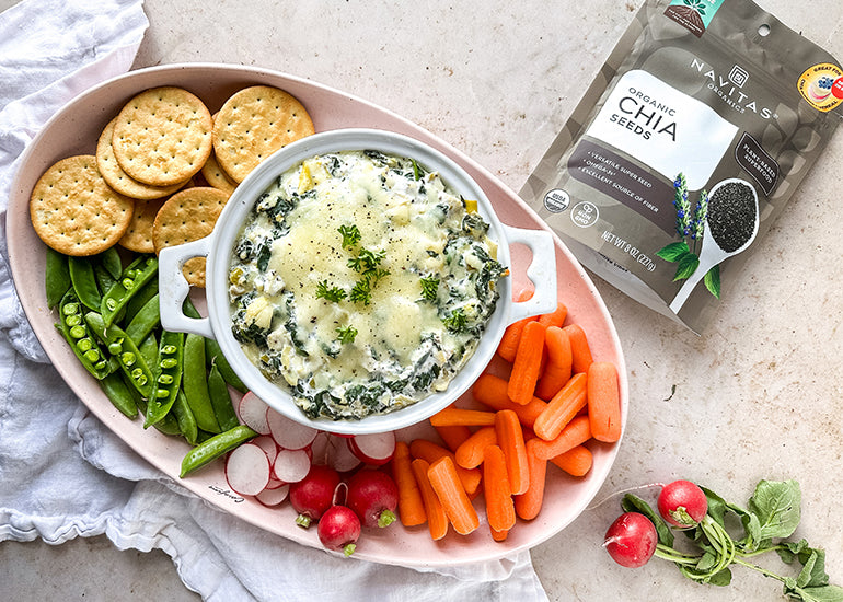 A platter filled with fresh vegetables and crackers surrounding a dish of spinach artichoke dip made with Navitas Organics Chia Seeds