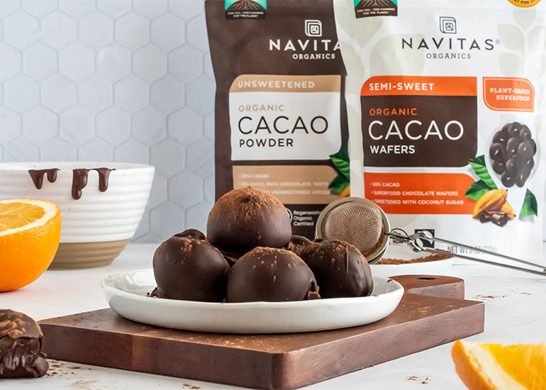 A wooden cutting board topped with a plate filled with chocolate orange truffles prepared with Navitas Organics Semi-sweet Cacao Wafers, sprinkled with Navitas Organics Cacao Powder