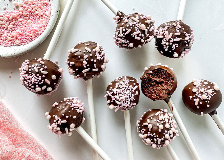 Nine chocolate cake pops made with Navitas Organics Cacao Powder, decorated with pink sprinkles, laying flat on a white surface