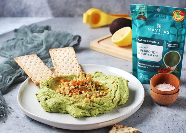 A plate topped with avocado hummus made with Navitas Organics Superfood+ Sea Veggie Blend, garnished with olive oil, paprika and pine nuts, with two crackers dipped into it
