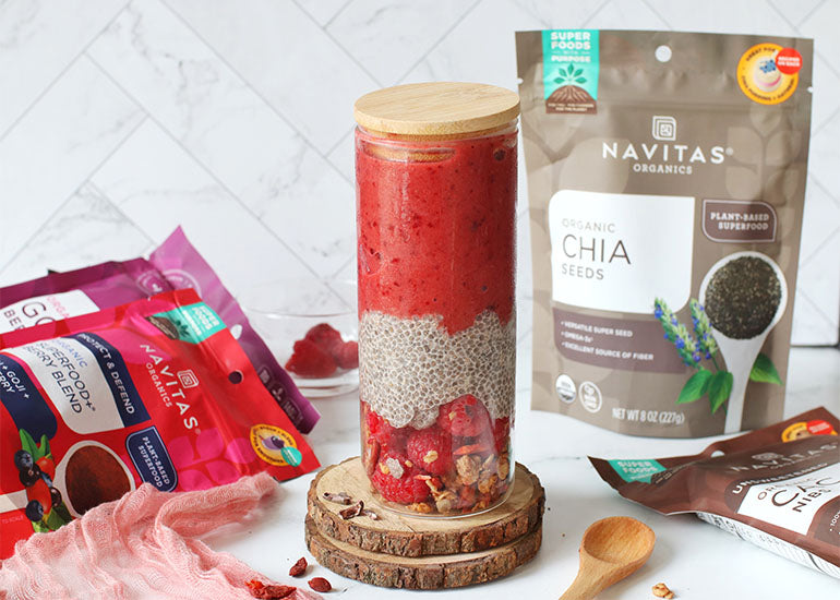 A tall chia pudding smoothie parfait in a glass surrounded by bags of Navitas Organics Superfood+ Berry Blend, Goji Berries and Chia Seeds