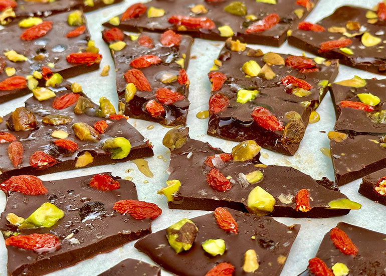 Spread out pieces of chocolate bark made with Navitas Organics Bittersweet Cacao Wafers topped with crushed pistachios and Navitas Organics Goji Berries and drizzled with maple syrup