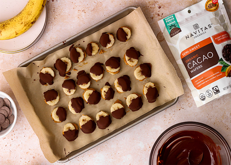 A parchment-lined pan topped with rows of chocolate-covered frozen banana bites made with Navitas Organics Semi-sweet Cacao Wafers