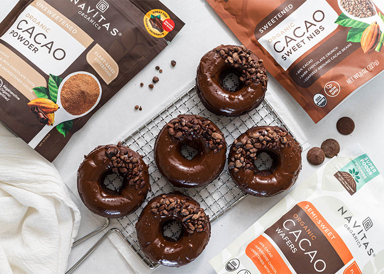 A cooling rack topped with five chocolate donuts made with Navitas Organics Cacao Powder, coated in a glaze made with Navitas Organics Semi-sweet Cacao Wafers and sprinkled with Navitas Organics Cacao Sweet Nibs