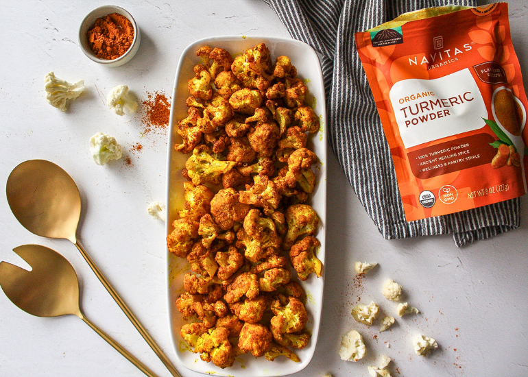 A long white dish topped with roasted turmeric cauliflower next to a bag of Navitas Organics Turmeric Powder, a small dish filled with turmeric powder and gold serving utensils