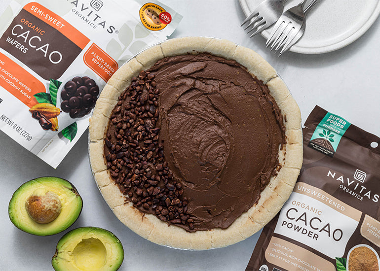 A prepared cacao avocado pie made with Navitas Organics Regnerative Organic Certified Cacao Powder and organic Semi-sweet Cacao Wafers surrounded by bags of product and a fresh avocado