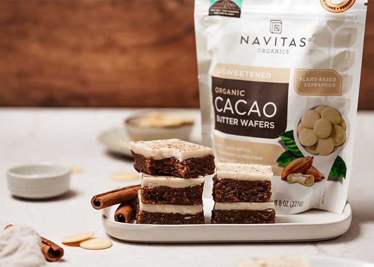 A stack of no-bake gingerbread cookie dough bars on a white plate in front of a bag of Navitas Organics Cacao Butter Wafers