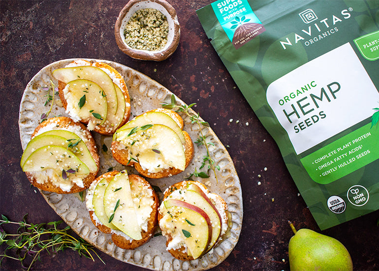 A decorative platter topped with pear and goat cheese crostinis beside a bag and small dish of Navitas Organics Hemp Seeds.