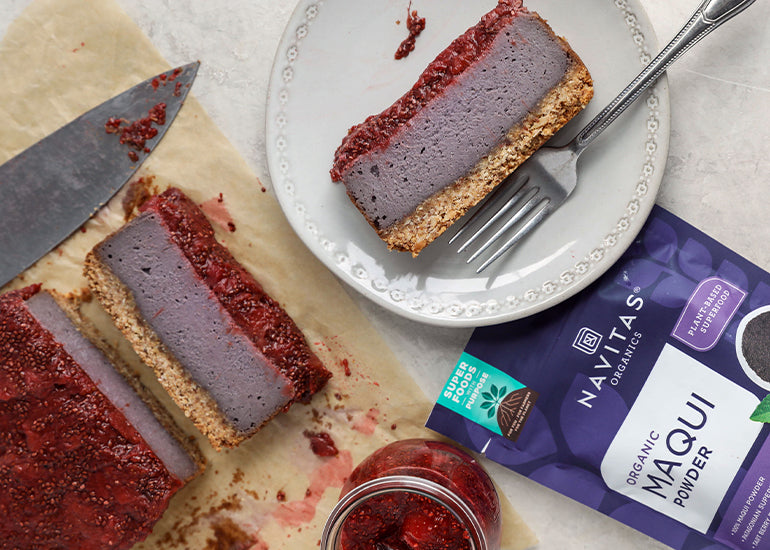 A plate topped with a slice of maqui berry cheesecake surrounded by a jar of strawberry topping and a bag of Navitas Organics Maqui Powder.