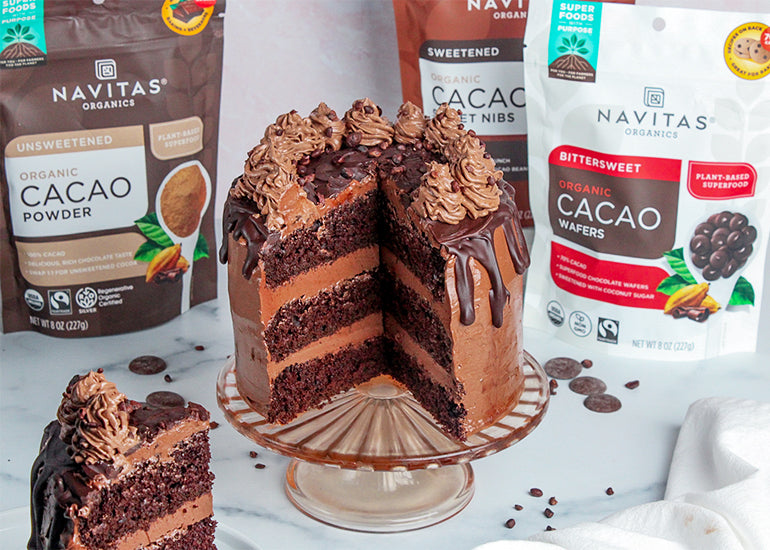 A triple chocolate cake sitting on a cake platter surrounded by bags of Navitas Organics Cacao Powder, Cacao Sweet Nibs and Bittersweet Cacao Wafers.