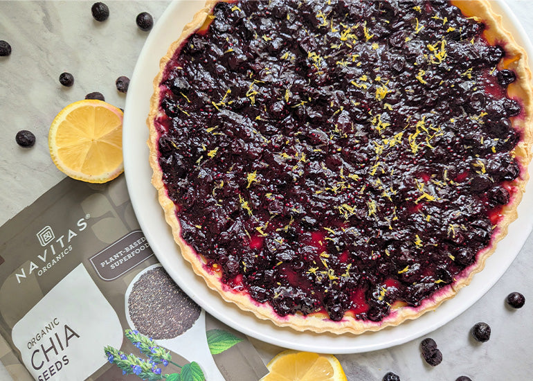 A plate with a lemon blueberry chia seed tart next to scattered blueberries and halved lemons with a bag of Navitas Organics Chia Seeds laying in the background.