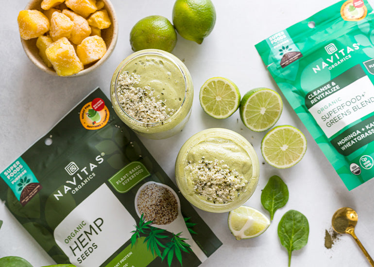 An overhead shot of two green smoothies topped with hemp seeds surrounded by a dish of frozen mango, halved limes, mint leaves, and bags of Navitas Organics Hemp Seeds and Superfood+ Greens Blend.