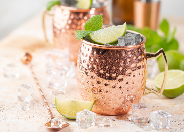 Two moscow mule cocktails on a counter garnished with lime wedges and mint leaves, surrounded by ice cubes and a stirring spoon.