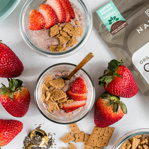 An overhead view of two jars of strawberry chia pudding, topped with crush graham crackers and sliced strawberries, surrounded by fresh strawberries and a bag of Navitas Organics Chia Seeds.