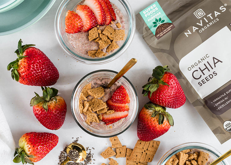 An overhead view of two jars of strawberry chia pudding, topped with crush graham crackers and sliced strawberries, surrounded by fresh strawberries and a bag of Navitas Organics Chia Seeds.