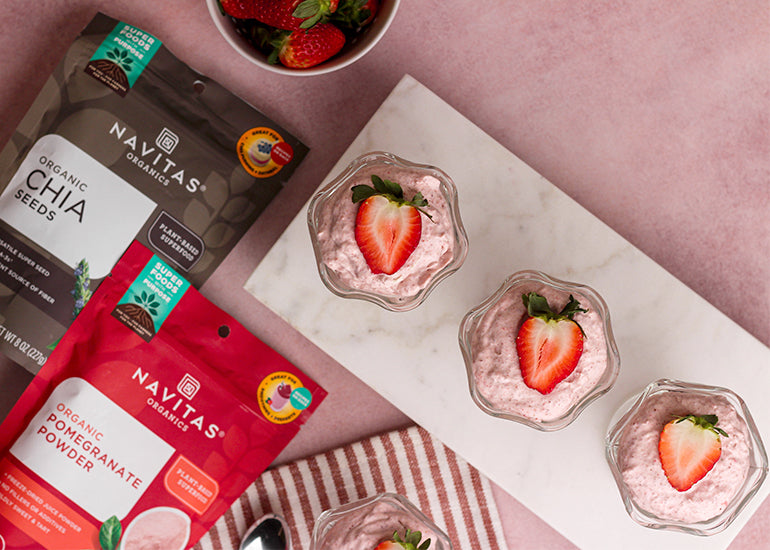 An overhead shot of three strawberry mousse cups topped with fresh strawberries lined up on a marble cutting board next to a bowl of fresh strawberries, and bags of Navitas Organics Chia Seeds and Pomegranate Powder.