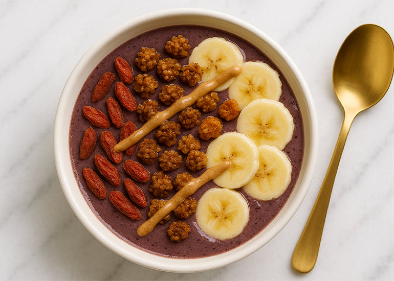 An acai smoothie bowl topped with Navitas Organics Goji Berries, Mulberries and Goldenberries, drizzled with nut butter and topped with banana slices next to a gold spoon on a white marble counter.
