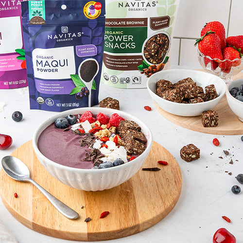 A maqui berry smoothie bowl topped with fruit, nuts and seeds, accompanied by two wooden plates holding fresh berries and chocolate snacks. In the background are bags of Navitas Organics Maqui Powder and Chocolate Brownie Power Snacks. 