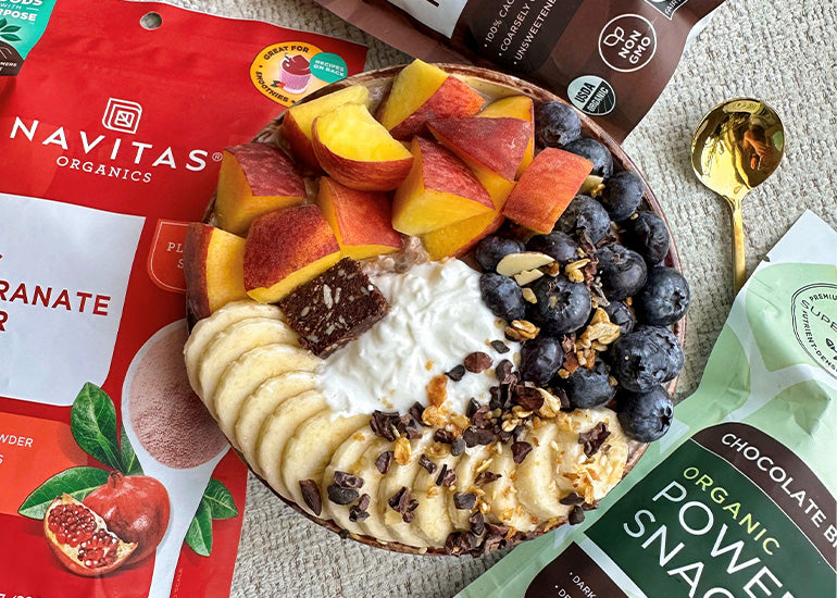 A healthy oatmeal bowl topped with various fruits like blueberries, banana slices and nectarines, accompanied by bags of Navitas Organics Pomegranate Powder, Cacao Nibs and Chocolate Brownie Power Snacks and a gold spoon.