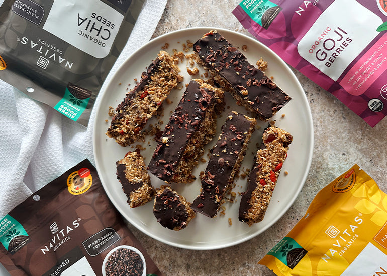 A plate of chocolate-covered granola bars surrounded by various packages of Navitas Organics products, including Cacao Nibs, Goji Berries, Goldenberries and Chia Seeds.