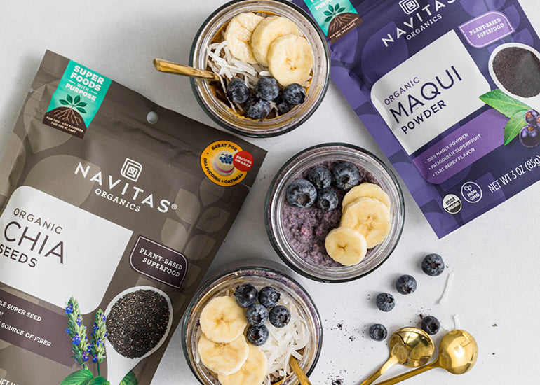 Overhead view of two bowls of maqui chia pudding topped with bananas, blueberries, and coconut, next to bags of Navitas Organics Chia Seeds and Maqui Powder.