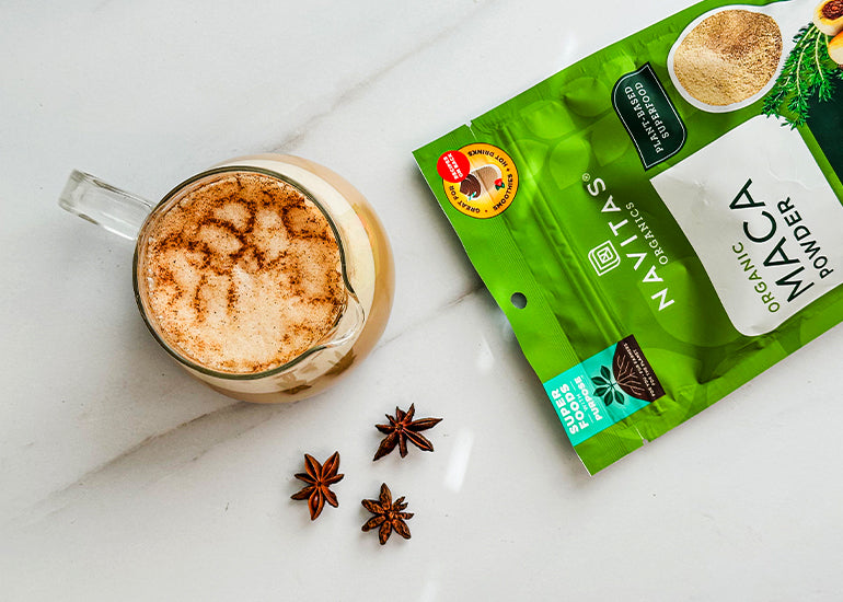 A glass mug filled with a creamy latte dusted with cinnamon next to a bag of Navitas Organics Maca Powder on a marble surface, accompanied by star anise.