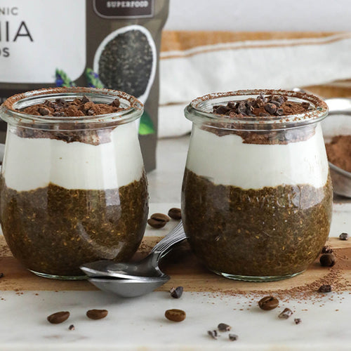 Two jars of layered chia pudding on a marble countertop, next to a bag of Navitas Organics Chia Seeds and scattered coffee beans around. Each jar is topped with white cream and Navitas Organics Cacao Nibs, accompanied by a spoon on the side.