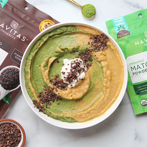 A bowl of swirled green and orange smoothie bowl topped with coconut cream and cacao nibs, surrounded by packages of Navitas Organics Cacao Nibs and Matcha Powder.